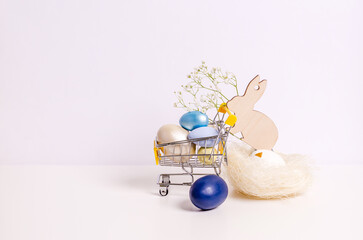 Easter wooden hare with a sprig of hepsophila carries a shopping cart with colored eggs on a white background.