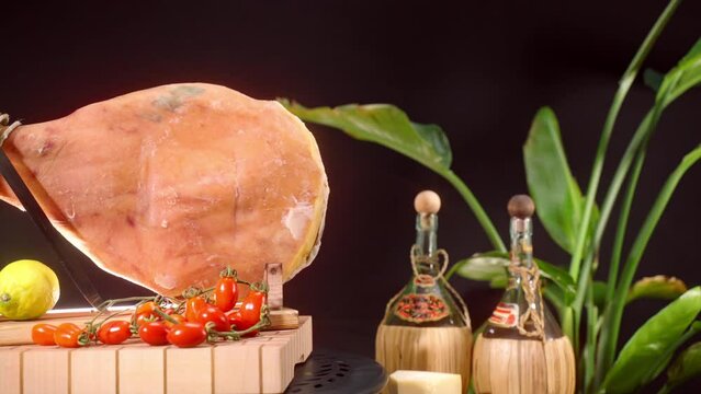 Dry cured ham on wooden stand with knife for slicing, lemon and fresh tomatoes against black background, salted jamon leg as an appetising antipasto of Italian and Spanish cuisine, typical cured