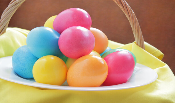 Beautiful, bright, multi-colored Easter eggs in a wooden basket on a white plate, decorated with yellow cloth. Easter eggs are found on a wooden background.
