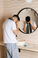 The young man is wrapped in a scarf and is sick with a cold, with a red face from the illness. Feeling unwell, flu, fever. Looking in the mirror in a bathroom.