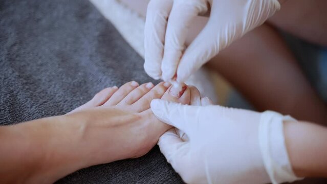 Toenail procedure in beauty salon, cosmetologist in gloves removing old nail polish with cotton pad and preparing woman foot for soaking in warm soap water. Chiropodist taking care of client's feet