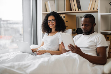 Handsome bearded man making notes on tablet while smiling bespectacled woman activating computer on her knees. Multiracial married couple doing efficient distant work while staying in bed at home.