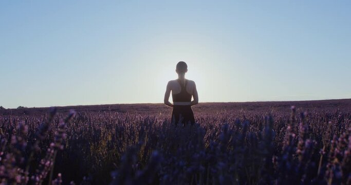 Female blogger practices yoga sun salutation asanas in pink and purple lavender fields at sunrise. Dreamy and inspiring morning. Free and happy woman stretching in sports wear. Meditational concept. 
