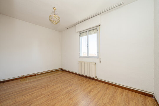 An empty room with ugly wooden floors, water pipes on the outside of the walls and an aluminum window