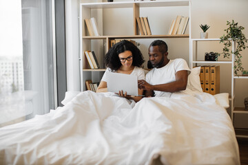 Fototapeta premium Beautiful young woman sitting in bed and sending text messages using modern laptop while her husband assisting her with advice. Loving married couple achieving tech-life balance resting at home.