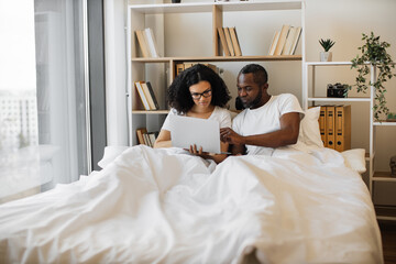 Obraz na płótnie Canvas Beautiful young woman sitting in bed and sending text messages using modern laptop while her husband assisting her with advice. Loving married couple achieving tech-life balance resting at home.