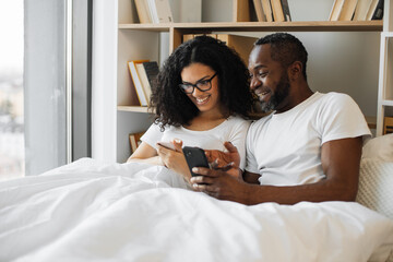 Serene multiracial family of two using mobile phones while lazing in soft bed during daytime at home. Confident adult man and bespectacled woman reading online article paying attention to details.