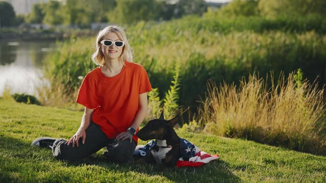 Portrait of the Beautiful Stylish Woman Sitting on the Grass with the Mini Bull Terrier Is Wrapped in the USA Flag in the Park. Patriotic Dog Lying on the Grass Near Girl Looking at Camera