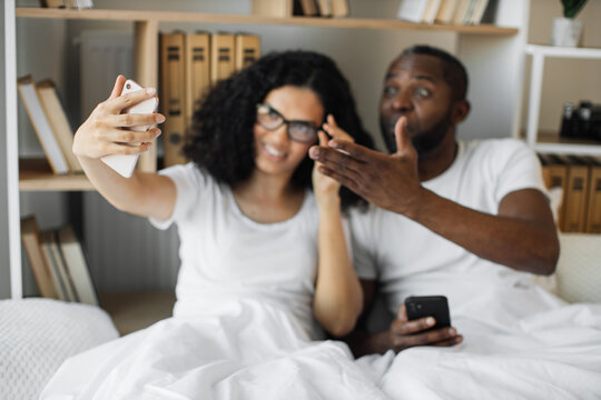 Focus on modern smartphone taking selfie of beautiful woman in glasses and surprised man with mobile lying in bed at home. Happy african american couple sharing experience of comfortable lifestyle.