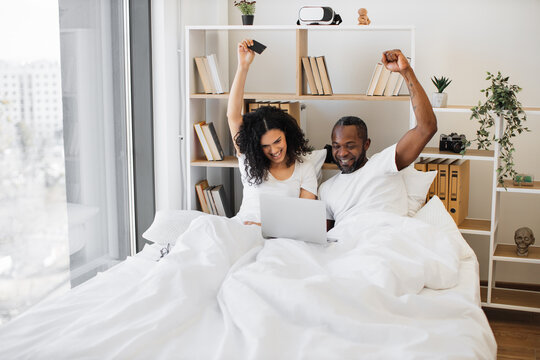 Emotional african american family of two raising hands in excitement while sitting in bed with laptop on blanket and credit card in hand. Thrilled married couple making successful deal online at home. - Powered by Adobe