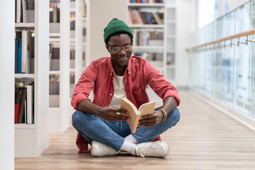 Happy African American millennial guy wearing glasses sitting on floor with book in hands, visiting...