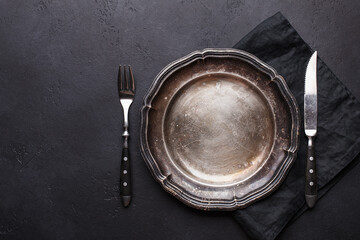 Empty vintage silver plate with fork, knife and linen napkin on black stone background. Top view...