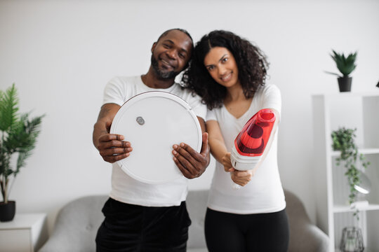 Smiling african man and woman in casual wear holding automatic vacuum cleaners while posing at living room. Concept of people, chores and modern technology.