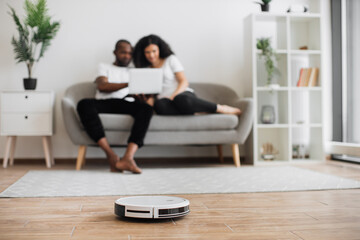 Obraz premium Automatic robot vacuum cleaning wooden floor while happy young couple resting on background. Two multicultural in love spending time for watching movie instead of households.