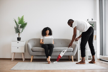 Side view of african man cleaning living room with cordless vacuum while focused woman working on laptop on couch. Caring husband doing households while wife freelancing at home.