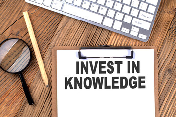 INVEST IN KNOWLEDGE text on paper clipboard with magnifier and keyboard on wooden background