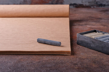 A dark gray drawing pastel crayon lies on a craft sketchbook next to a black pastel box. Selective focus. The concept of the artist's working corner, materials and tools for drawing