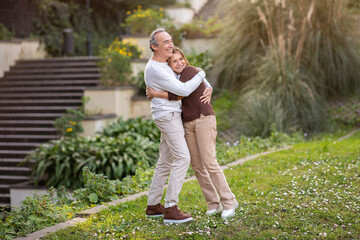 Mature Spouses Embracing Standing In Green Park Outside, Looking Aside