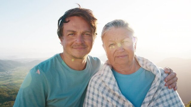 Portrait of young man and elderly woman. Young man hugs senior caucasian woman outdoor at sunrise. Models look into the camera