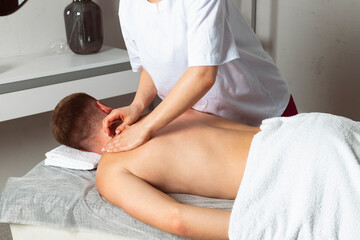 Fototapeta na wymiar A professional masseuse or massage therapist is massaging the client's shoulder blades while the client lies partially covered with a towel on the massage table. Concept of relaxation and self-care.