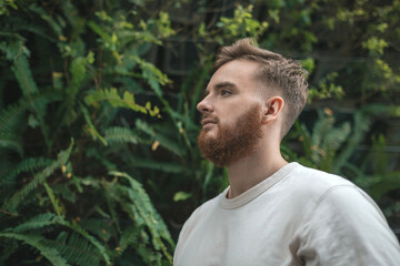 Naklejka na ściany i meble Portrait of happy young bearded man with beard breathing deep, inhale fresh air on natural background, green wall with leaves, flower in tropical country in garden. Eco, ecological concept. 