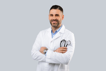 Confident male medical practitioner in white coat holding stethoscope in hand, posing with folded arms