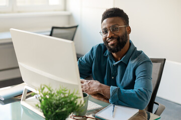 Positive black businessman in eyeglasses smiling at camera, sitting in front of computer at workdesk in office