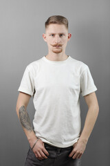 Handsome young man with stylish haircut, beard and handlebar moustache with tattooed arm in mock up white t shirt is looking at camera, hands in pockets isolated on grey background. Fashion concept