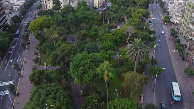 Public park called "Parque Kennedy" in between two streets, filled with many green trees. Drone flies forward while tilting gimbal camera down. Located in Miraflores district of Lima, Peru.