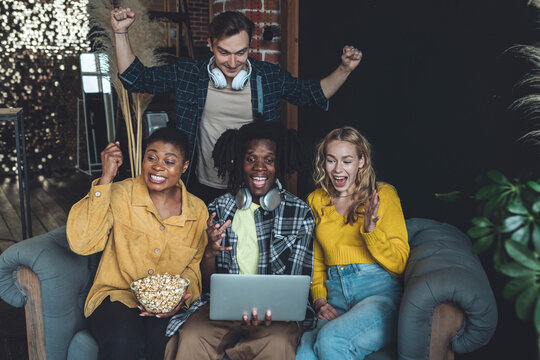 Diverse group of college students, friends, young people watching football or baseball game on laptop, have fun, eating popcorn. Concept of international friendship, leisure time, home activity