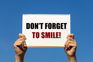 Don't forget to smile text on notebook paper held by 2 hands with isolated blue sky background. This message can be used as business concept about being happy.