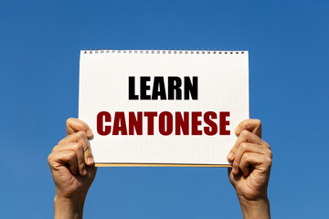 Learn Cantonese text on notebook paper held by 2 hands with isolated blue sky background. This message can be used as business concept about learning Cantonese language.