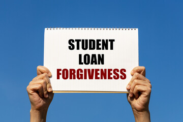 Student loan forgiveness text on notebook paper held by 2 hands with isolated blue sky background....