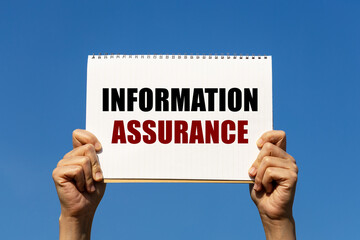 Information assurance text on notebook paper held by 2 hands with isolated blue sky background. This message can be used as business concept about information assurance.