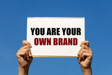 You are your own brand text on notebook paper held by 2 hands with isolated blue sky background. This message can be used as business concept about branding yourself.