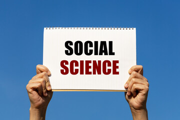 Social science text on notebook paper held by 2 hands with isolated blue sky background. This message can be used as business concept about social science.