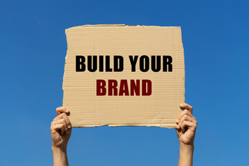 Build your brand text on box paper held by 2 hands with isolated blue sky background. This message board can be used as business concept about building your brand.