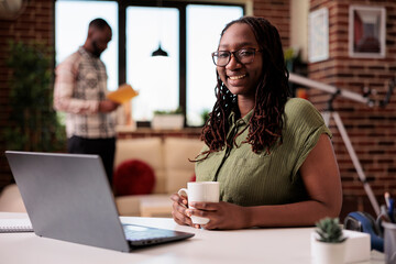 Portrait of african american freelancer smiling at camera while holding a cup of coffee or tea in home living room. Casual entrepreneur taking a break from remote work while roommate is reading a book