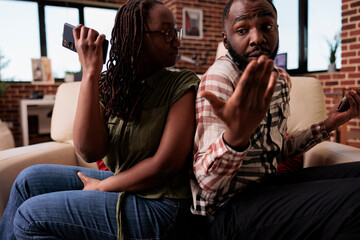 Woman threatening confused boyfriend about contents of smartphone arguing sitting back to back on...