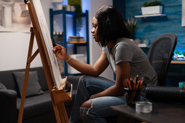 Artistic woman creating pencil sketch on easel at art workplace. African american female artist making hand drawing on canvas, sitting at home made sketching atelier in bedroom.