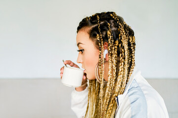 A young woman drinking coffee is in profile with braids in all her hair wearing a very large golden ring and headphones with a white and blue jacket