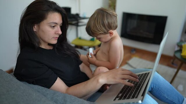 Parent multi tasking in front of laptop computer laying on couch. Toddler baby sitting on mother lap while working from home. Cute modern parenting lifestyle