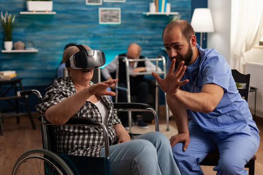Male Nurse Teaching Elderly Female Patient To Use Virtual Reality Goggles In Nursing Home Waiting Room. Woman In Wheelchair Having A Fun Day With Modern Simulation Technology.