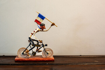 A small handmade french guy doll riding a bicycle and raising the French flag with his left arm, on an old wooden and iron trunk.