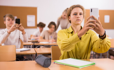 Teenage girl sitting at desk in classroom and photographing with her smartphone.