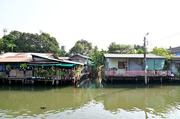 Fototapeta na wymiar BANGKOK, THAILAND - MARCH 30, 2023 : Wooden houses, Thai houses and buildings along the Chao Phraya River with white clouds and blue sky background, Town Bangkok Waterfront, Thailand.