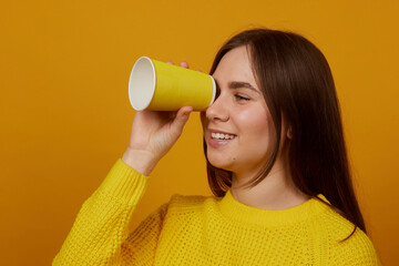 The girl holds a disposable glass in her hand and rejoices on a yellow background
