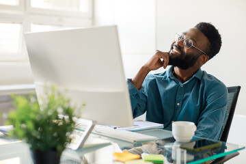 Optimistic dream. Happy black male employee sitting in front of computer and smiling, visualizing future career growth