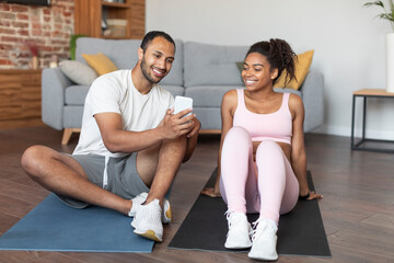 Glad young black guy in sportswear show video lesson on phone to female, sit on mat on floor