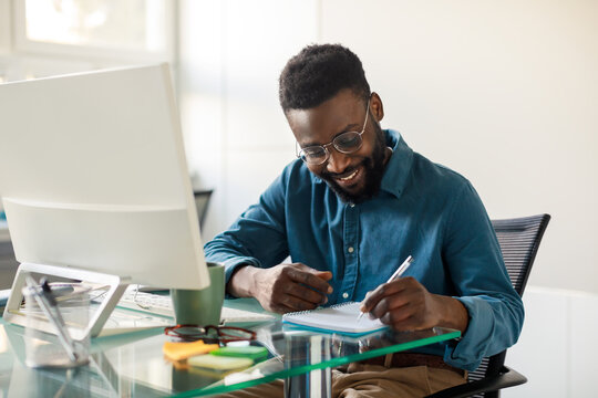 Positive black executive ceo manager writing notes in notebook, sitting in front of computer at workplace in office